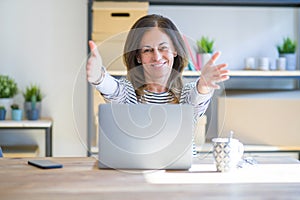 Middle age senior woman sitting at the table at home working using computer laptop looking at the camera smiling with open arms