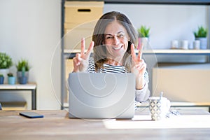 Middle age senior woman sitting at the table at home working using computer laptop smiling looking to the camera showing fingers