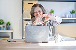 Middle age senior woman sitting at the table at home working using computer laptop smiling in love doing heart symbol shape with