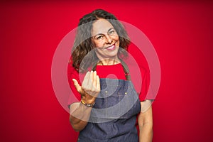 Middle age senior woman wearing apron uniform over red isolated background Beckoning come here gesture with hand inviting