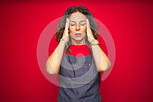 Middle age senior woman wearing apron uniform over red isolated background with hand on headache because stress