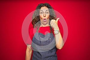 Middle age senior woman wearing apron uniform over red isolated background Surprised pointing with hand finger to the side, open