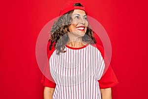 Middle age senior woman wearing baseball equiment over red isolated background looking away to side with smile on face, natural