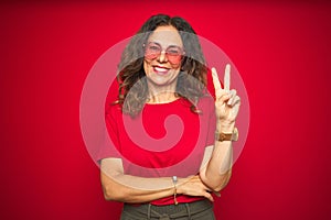 Middle age senior woman wearing cute heart shaped glasses over red isolated background smiling with happy face winking at the