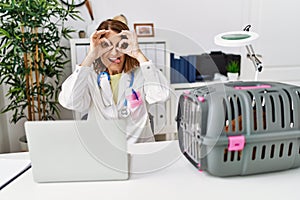 Middle age veterinarian woman working at pet clinic doing ok gesture like binoculars sticking tongue out, eyes looking through