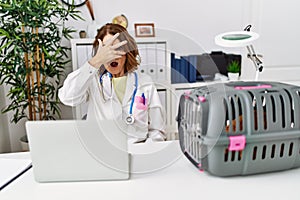 Middle age veterinarian woman working at pet clinic peeking in shock covering face and eyes with hand, looking through fingers