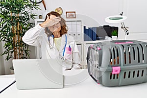 Middle age veterinarian woman working at pet clinic smiling and laughing with hand on face covering eyes for surprise