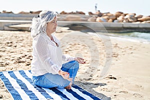 Middle age woman doing yoga exercise sitting on towel at seaside