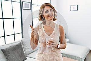 Middle age woman drinking a cup coffee at home doing happy thumbs up gesture with hand