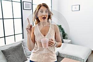 Middle age woman drinking a cup coffee at home pointing finger up with successful idea