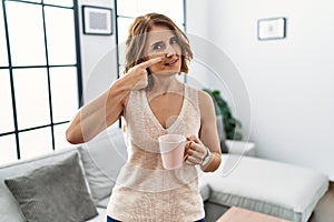 Middle age woman drinking a cup coffee at home pointing with hand finger to face and nose, smiling cheerful