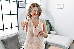 Middle age woman drinking a cup coffee at home pointing to you and the camera with fingers, smiling positive and cheerful