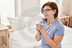 Middle age woman drinking cup of coffee sitting on bed at bedroom