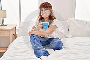 Middle age woman drinking cup of coffee sitting on bed at bedroom