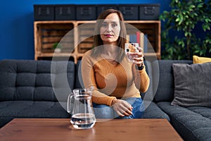 Middle age woman drinking glass of water thinking attitude and sober expression looking self confident