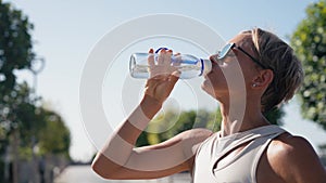 Middle age woman drinking water from plastic bottle after gym, outdoor workout, city sport, brunette caucasian girl