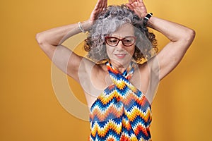 Middle age woman with grey hair standing over yellow background doing bunny ears gesture with hands palms looking cynical and