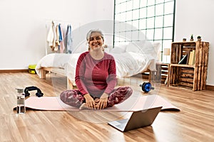 Middle age woman with grey hair training at home looking at exercise video on laptop looking positive and happy standing and