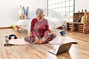 Middle age woman with grey hair training at home looking at exercise video on laptop scared and amazed with open mouth for