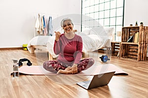 Middle age woman with grey hair training at home looking at exercise video on laptop smiling with happy face looking and pointing