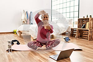 Middle age woman with grey hair training at home looking at exercise video on laptop smiling making frame with hands and fingers