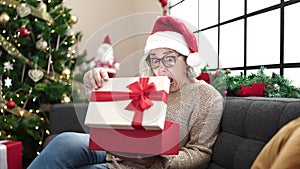 Middle age woman with grey hair wearing christmas hat unpacking gift with surprised face at home