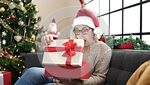 Middle age woman with grey hair wearing christmas hat unpacking gift with surprised face at home