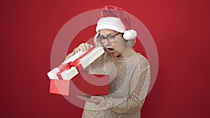 Middle age woman with grey hair wearing christmas hat unpacking gift with surprised face over isolated red background