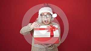 Middle age woman with grey hair wearing christmas hat unpacking gift with surprised face over isolated red background