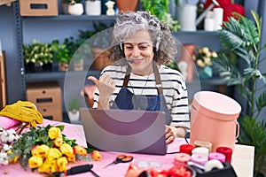Middle age woman with grey hair working at florist with laptop pointing thumb up to the side smiling happy with open mouth