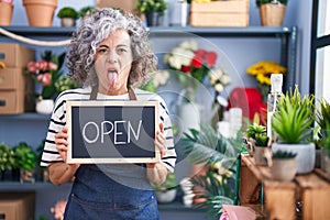 Middle age woman with grey hair working at florist with open sign sticking tongue out happy with funny expression