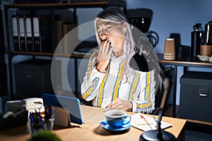Middle age woman with grey hair working at the office at night bored yawning tired covering mouth with hand
