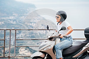 Middle-age woman in helmet and sunglasses on motor scooter on the Sicilian old town streets in the Forza d`Agro with Sant`Alessi