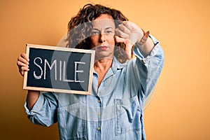 Middle age woman holding blackboard with smile message over isolated yellow background with angry face, negative sign showing