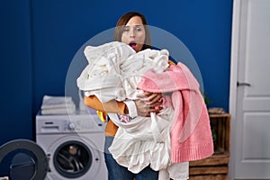 Middle age woman holding dirty laundry ready to put it in the washing machine afraid and shocked with surprise and amazed