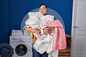 Middle age woman holding dirty laundry ready to put it in the washing machine puffing cheeks with funny face