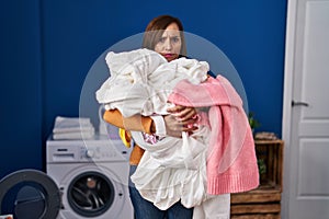 Middle age woman holding dirty laundry ready to put it in the washing machine skeptic and nervous, frowning upset because of