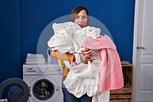 Middle age woman holding dirty laundry ready to put it in the washing machine skeptic and nervous, frowning upset because of