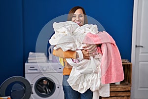 Middle age woman holding dirty laundry ready to put it in the washing machine smiling with a happy and cool smile on face