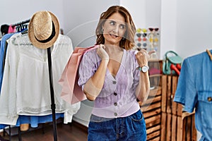 Middle age woman holding shopping bags at shop retail pointing thumb up to the side smiling happy with open mouth
