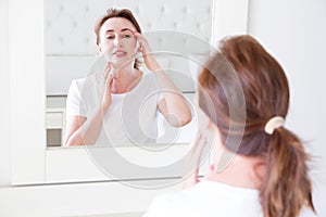 Middle age woman looking in mirror on face. Make up and lifting. Wrinkles and anti aging skin care concept. Selective focus