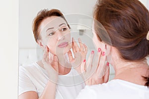 Middle age woman looking in mirror on face. Wrinkles and anti aging skin care concept. Selective focus