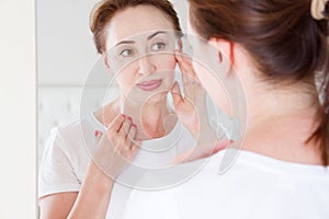 Middle age woman looking in mirror on face. Wrinkles and anti aging skin care concept. Selective focus