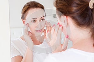 Middle age woman looking in mirror on face. Wrinkles and anti aging skin care concept. Selective focus