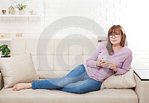 Middle age woman lying on sofa and using smartphone at home background. Copy space and mock up. Online shopping, internet