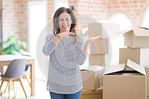 Middle age woman moving to a new house arround cardboard boxes amazed and smiling to the camera while presenting with hand and
