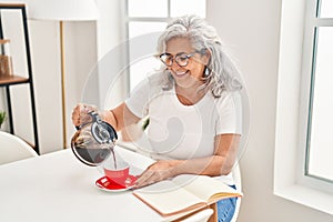 Middle age woman pouring coffee on cup sitting on table at home