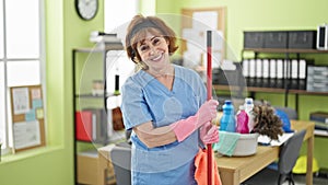 Middle age woman professional cleaner holding mop stick smiling at office