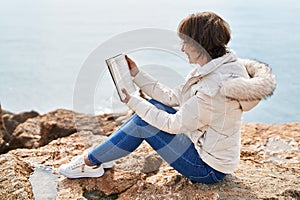 Middle age woman reading book sitting on the rock at seaside