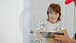 Middle age woman receiving package signing on clipboard at home